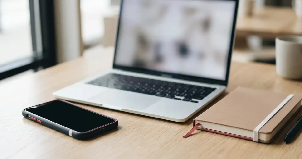 A smartphone on a table beside a laptop and a closed notebook